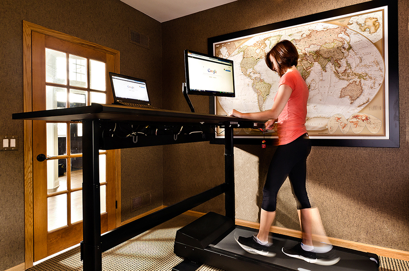 Dark haired woman walking on treadmill desk in front of global map framed art